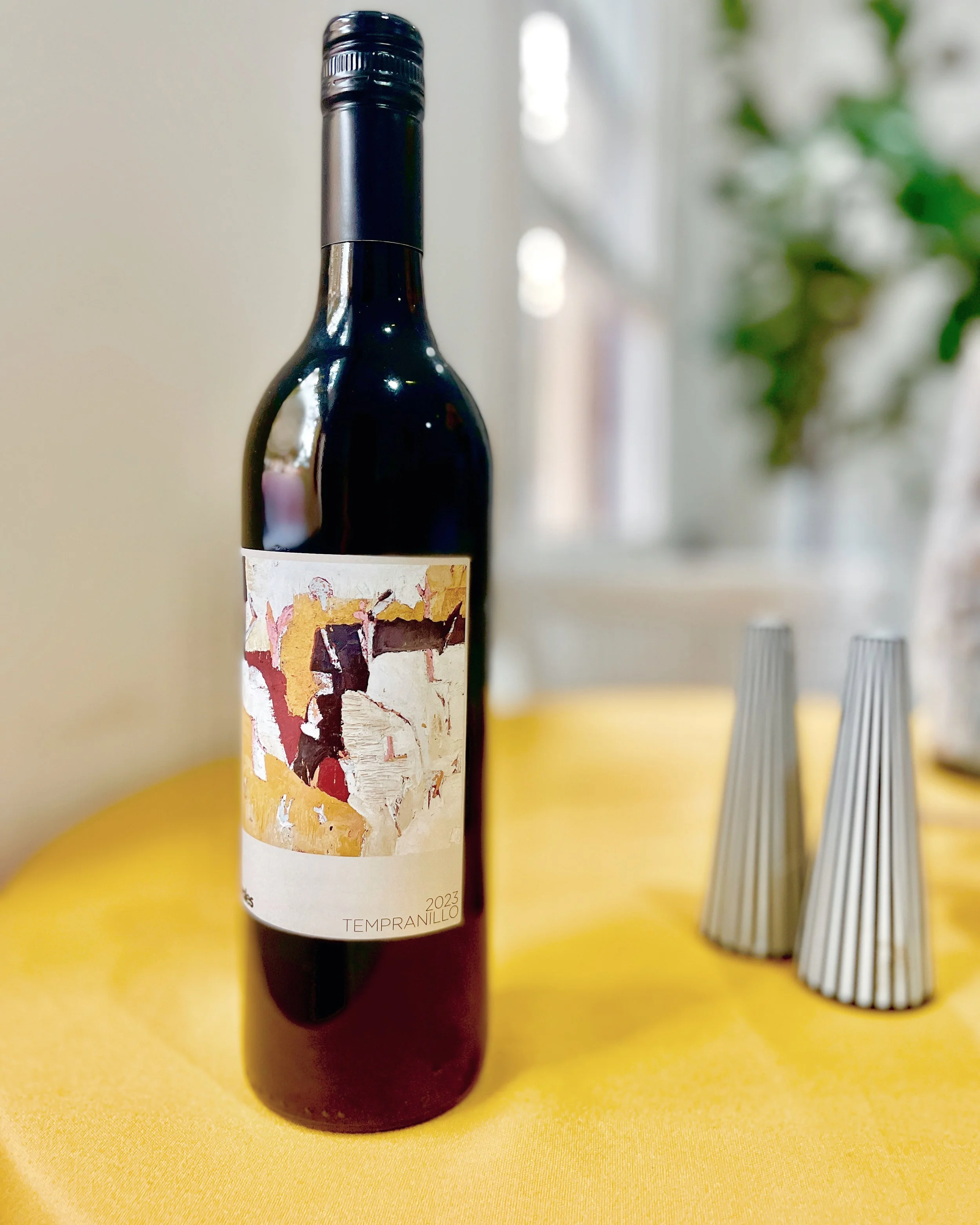 A bottle of Tempranillo red wine with an abstract art label, placed on a yellow tablecloth next to two metallic candle holders.