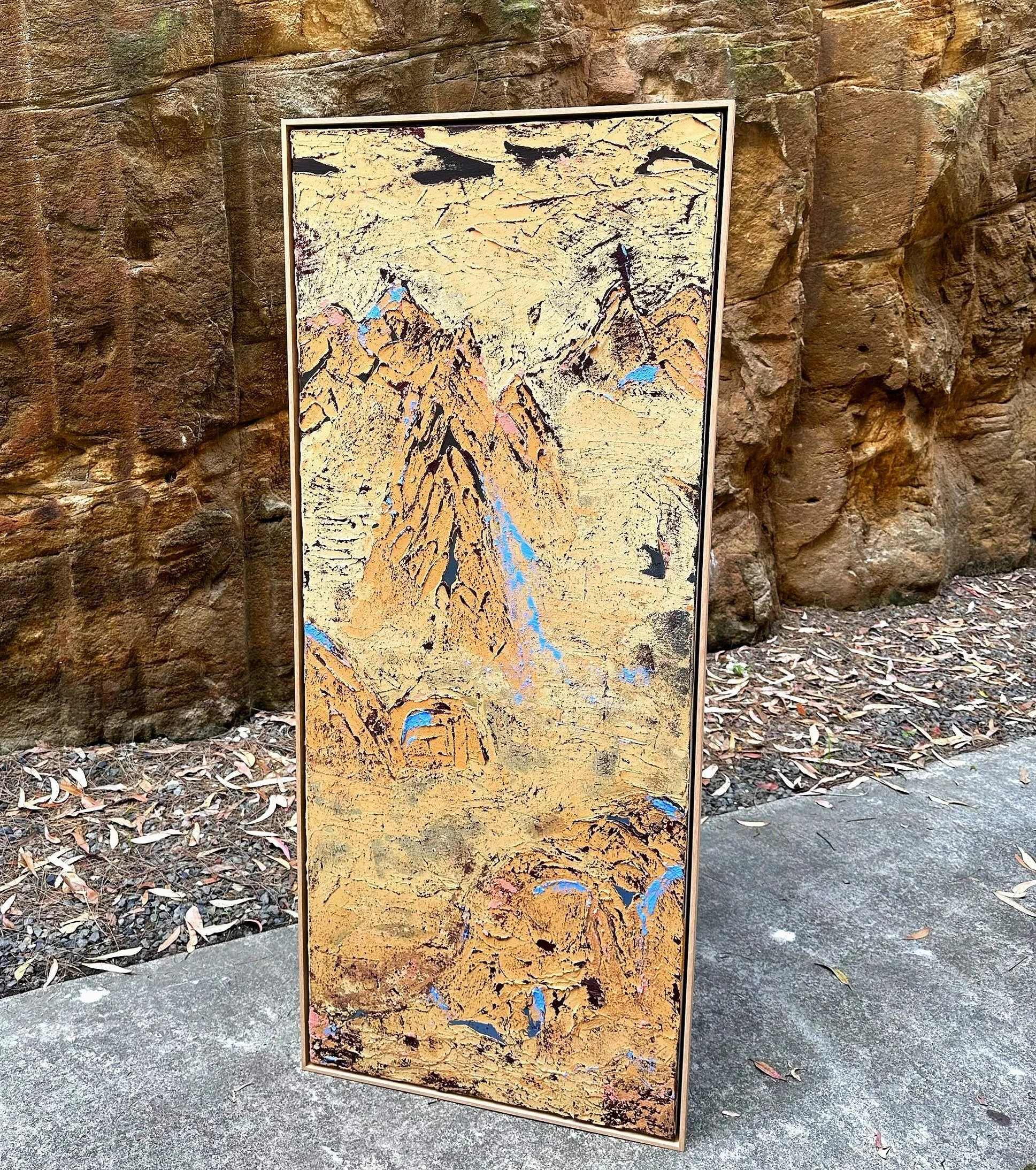An abstract painting with earthy tones, black lines, and touches of blue, displayed outdoors against a rocky wall.