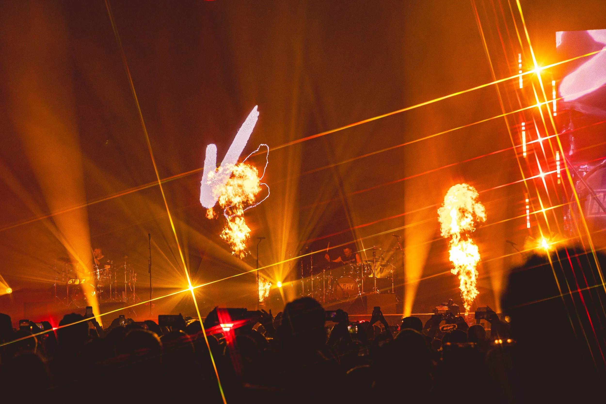 A concert stage with fire flames and pyrotechnics, bright yellow and red lights, and an audience taking photos, creating an energetic atmosphere.