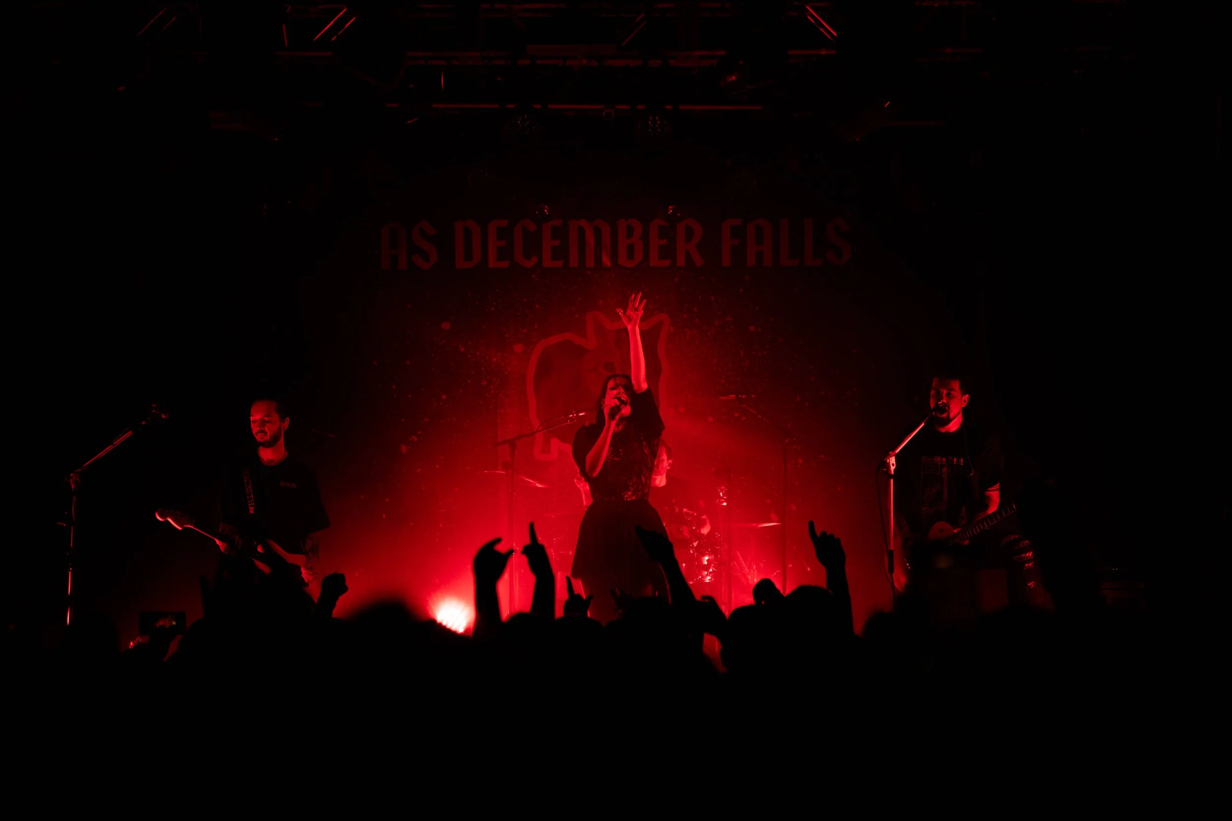 Band performing on stage with red lighting, audience in foreground with raised hands, background display reads 'As December Falls'
