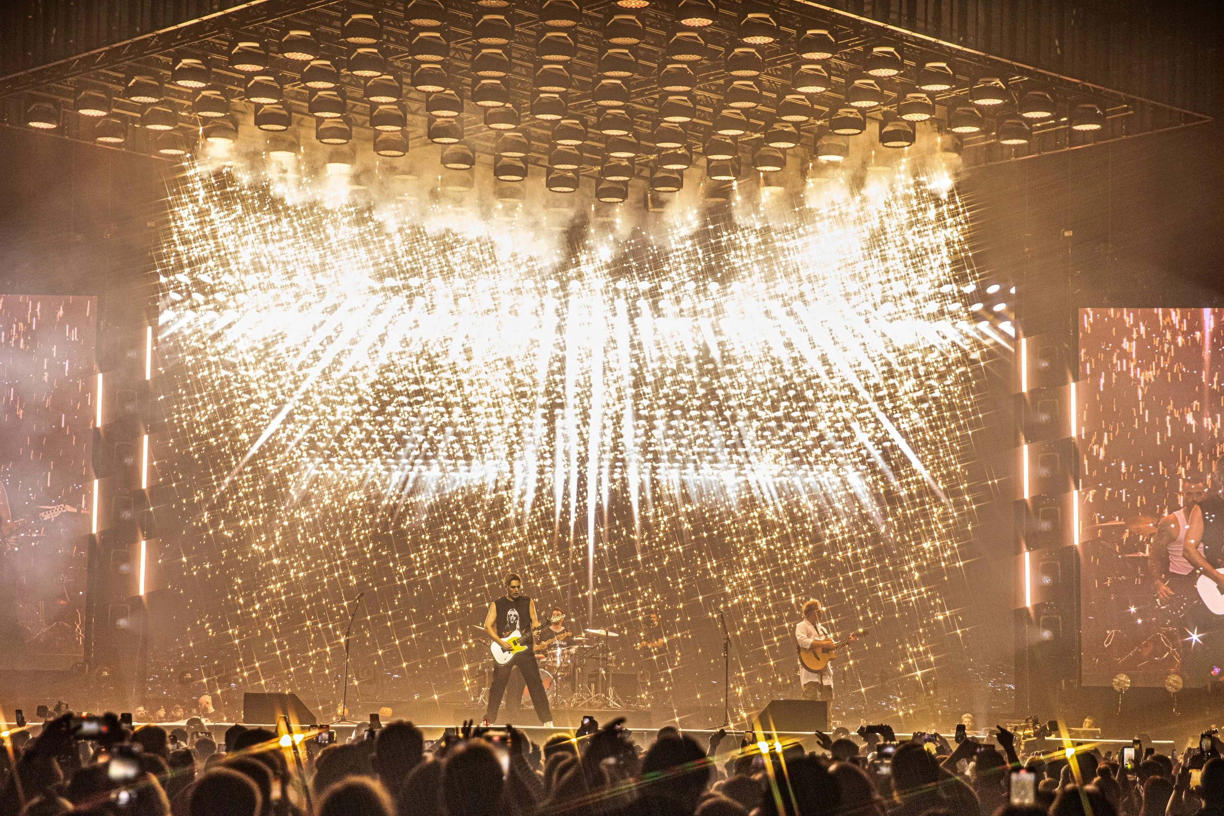 A live concert with a band playing on stage under a large, dazzling LED display with bright lights and sparkling visuals, and a large crowd of fans in the foreground.