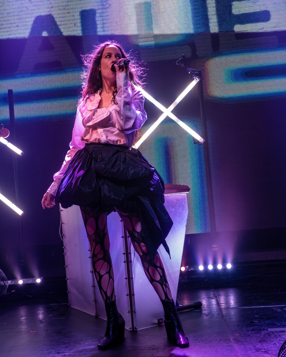 A female singer performing on stage with a microphone, wearing a pink blouse, a black skirt, and torn patterned stockings, with purple stage lighting and LED screens in the background.