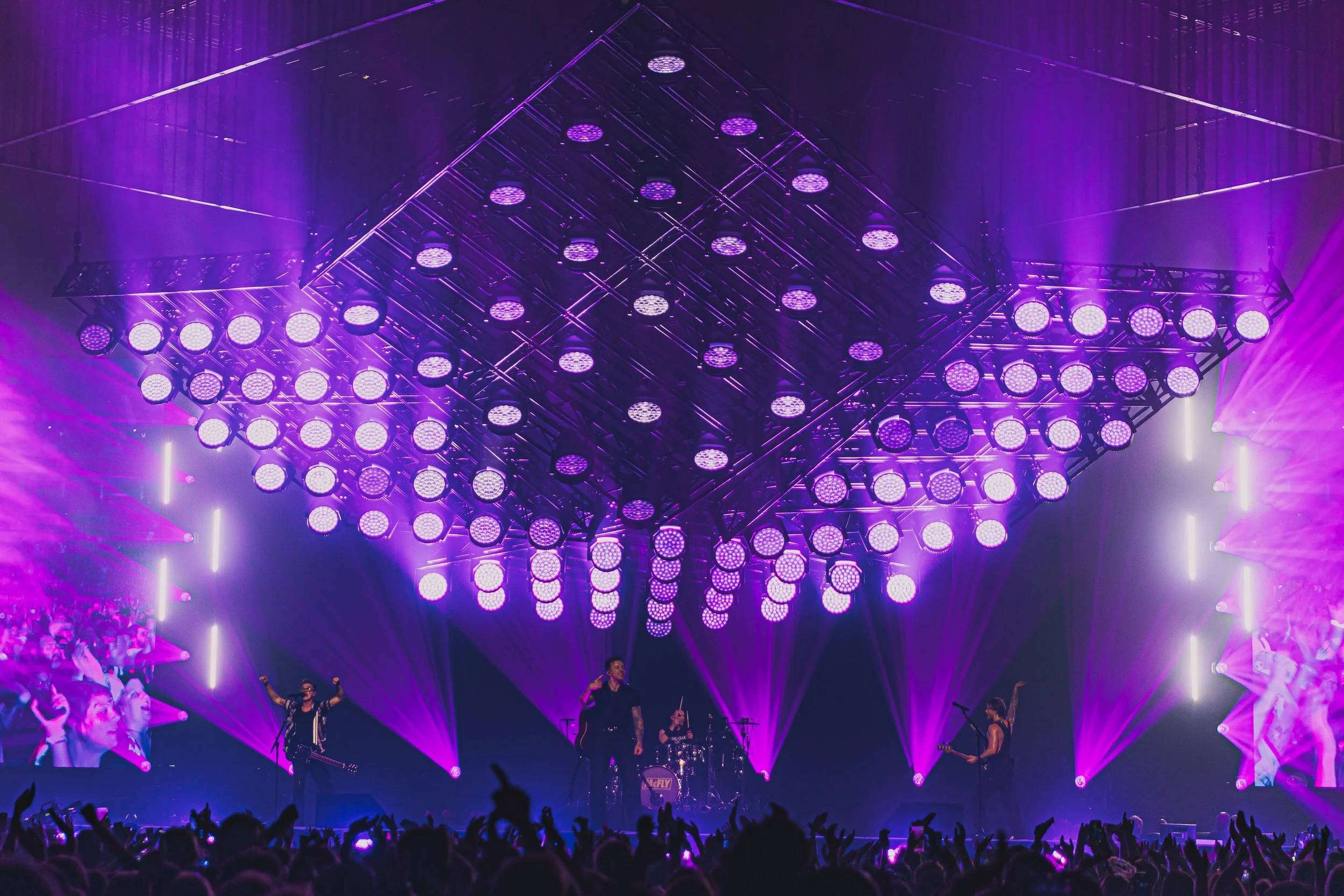 Concert stage with purple lighting, band performing, audience in foreground, large LED screens with visuals on sides.