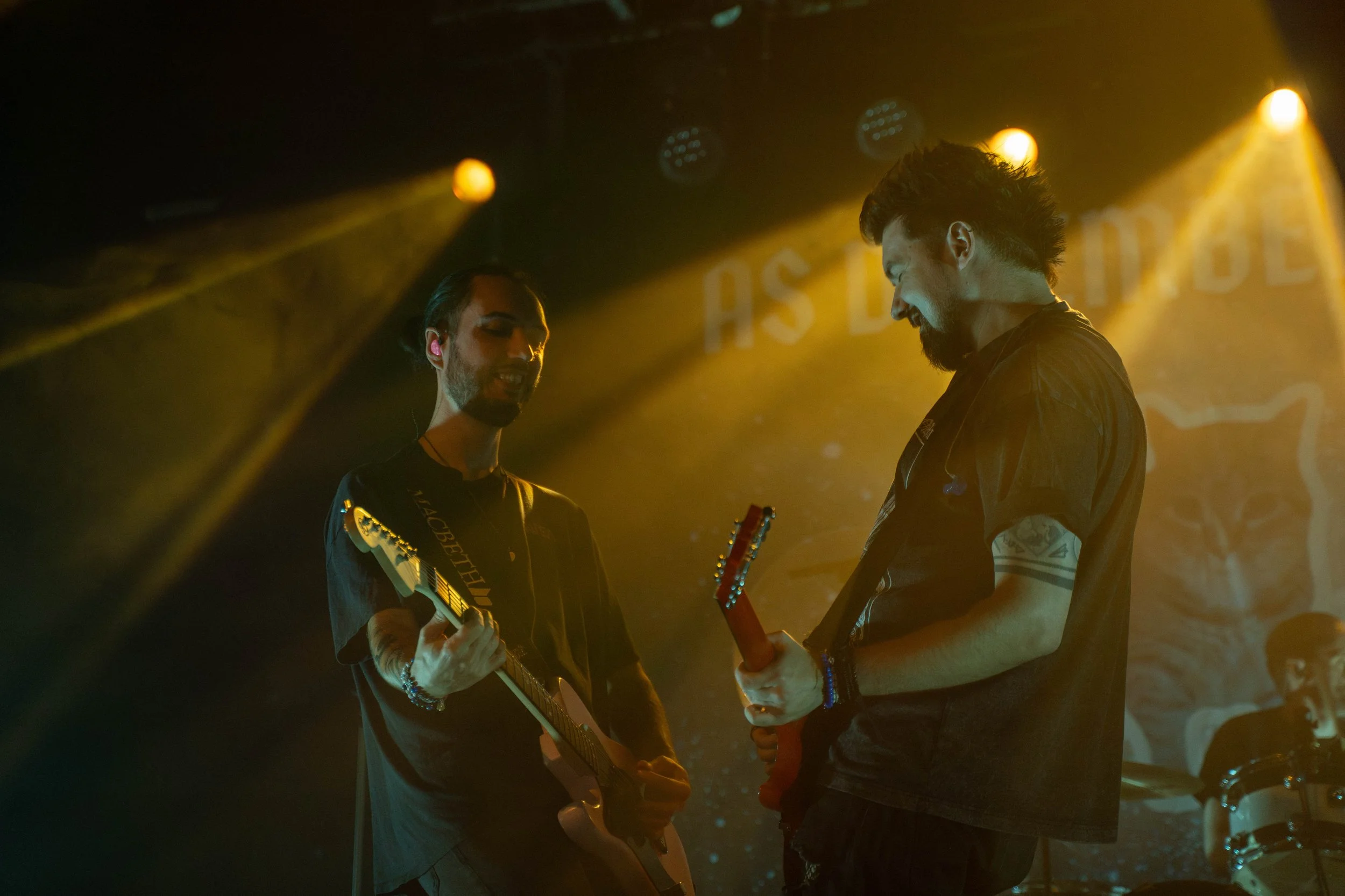 Two male musicians on stage playing electric guitars under yellow stage lights, with a drummer visible in the background and a partially visible backdrop with text.