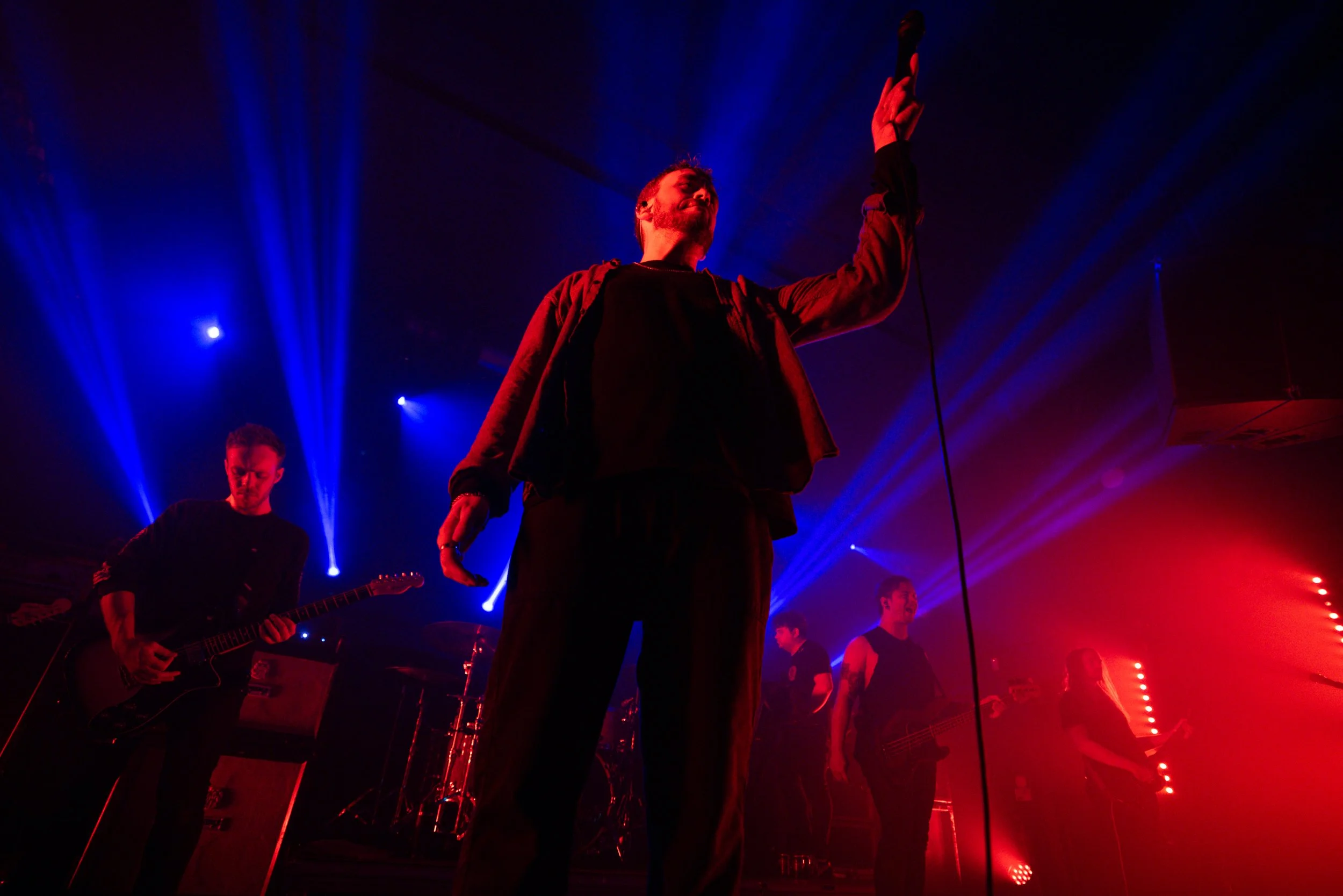 A band performs on stage with red and blue lighting. The lead singer is holding a microphone in the air, while the other band members play guitars and bass.