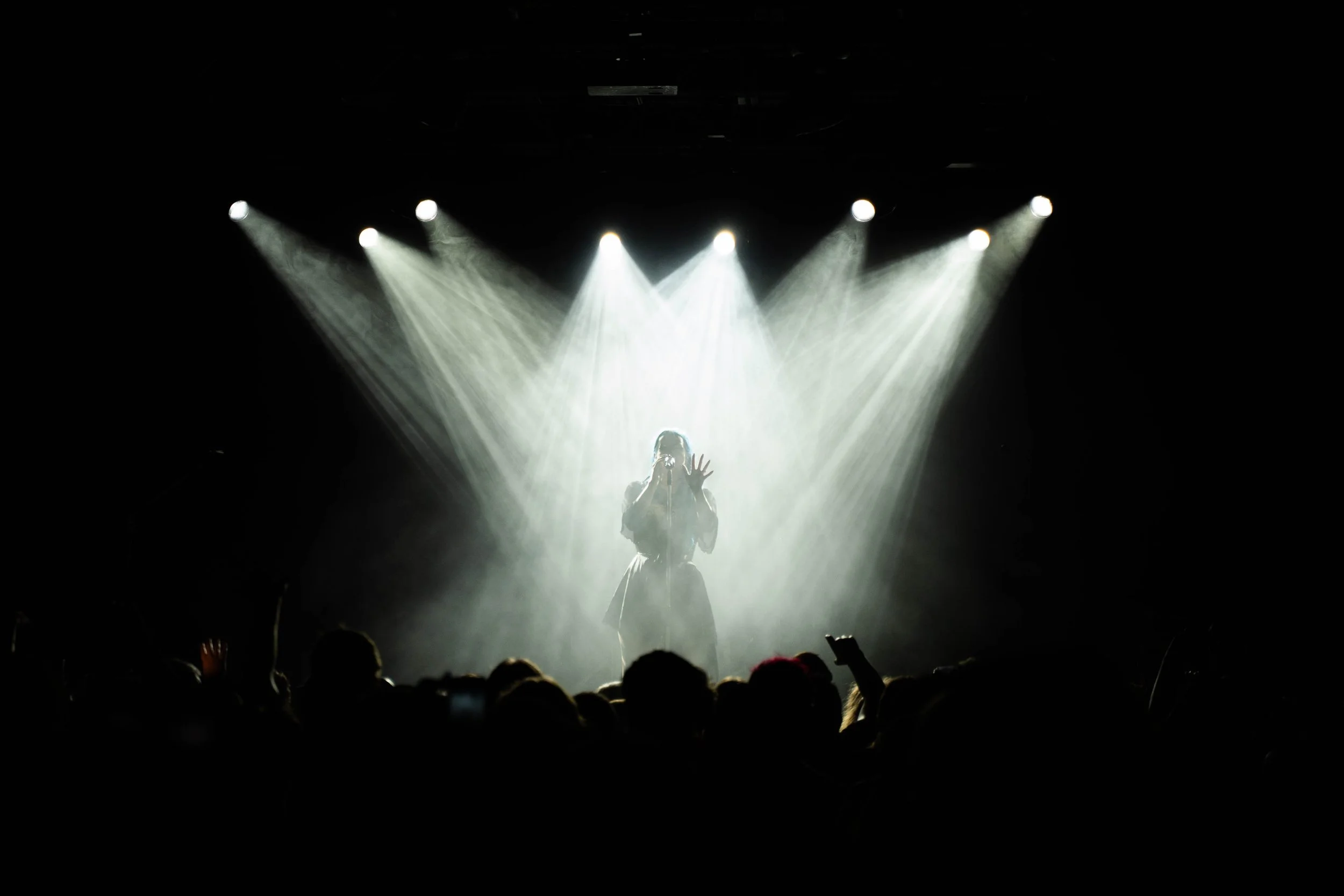 A performer on stage singing into a microphone with bright stage lights shining behind her, creating a dramatic atmosphere, with an audience visible in the foreground.
