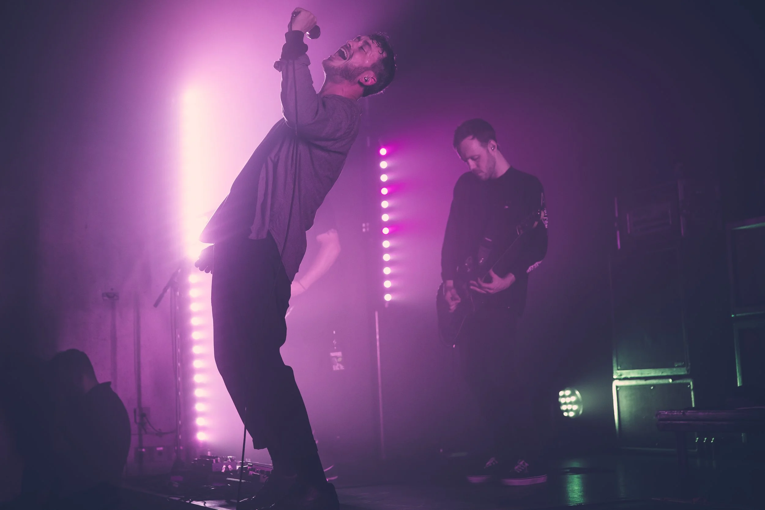 Two musicians performing on stage with vibrant purple lighting, one singing passionately into a microphone and the other playing an electric guitar.