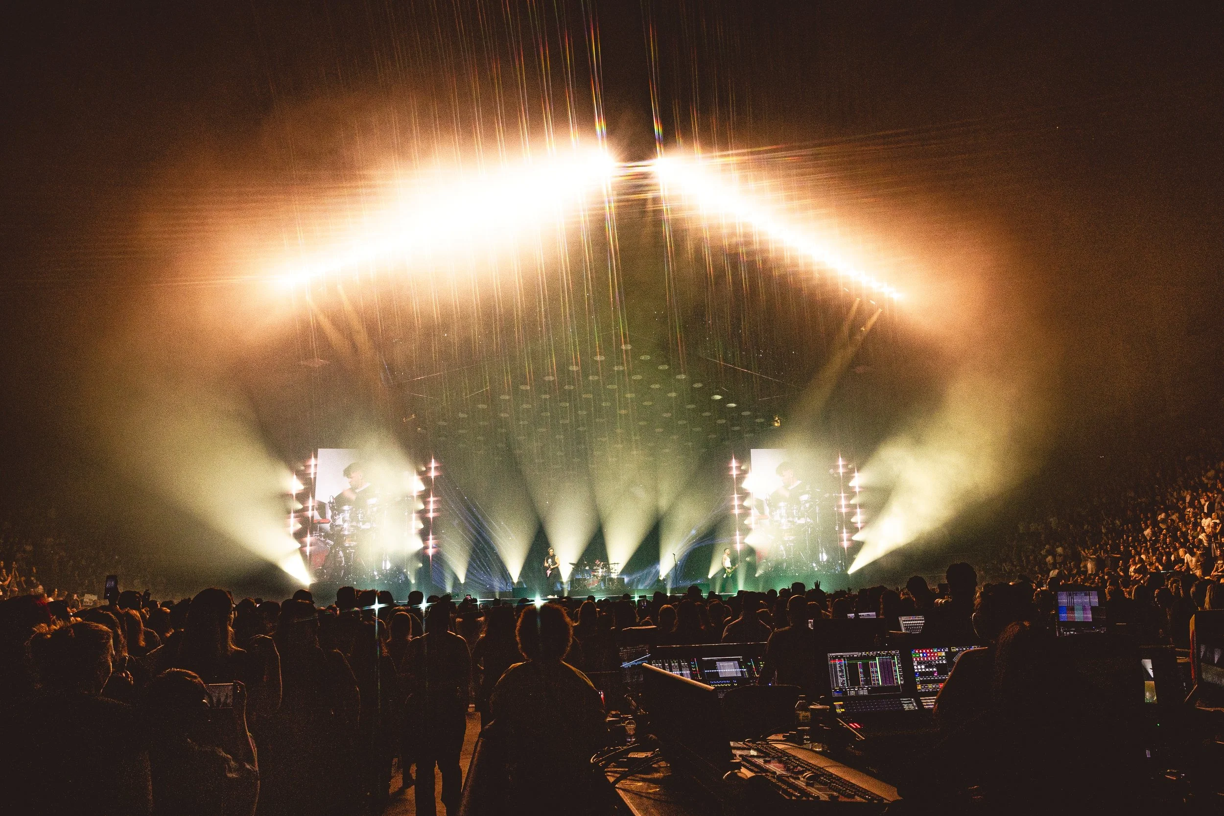 A concert stage with bright lights, large screens displaying musicians, and an audience watching in a dimly lit indoor arena.