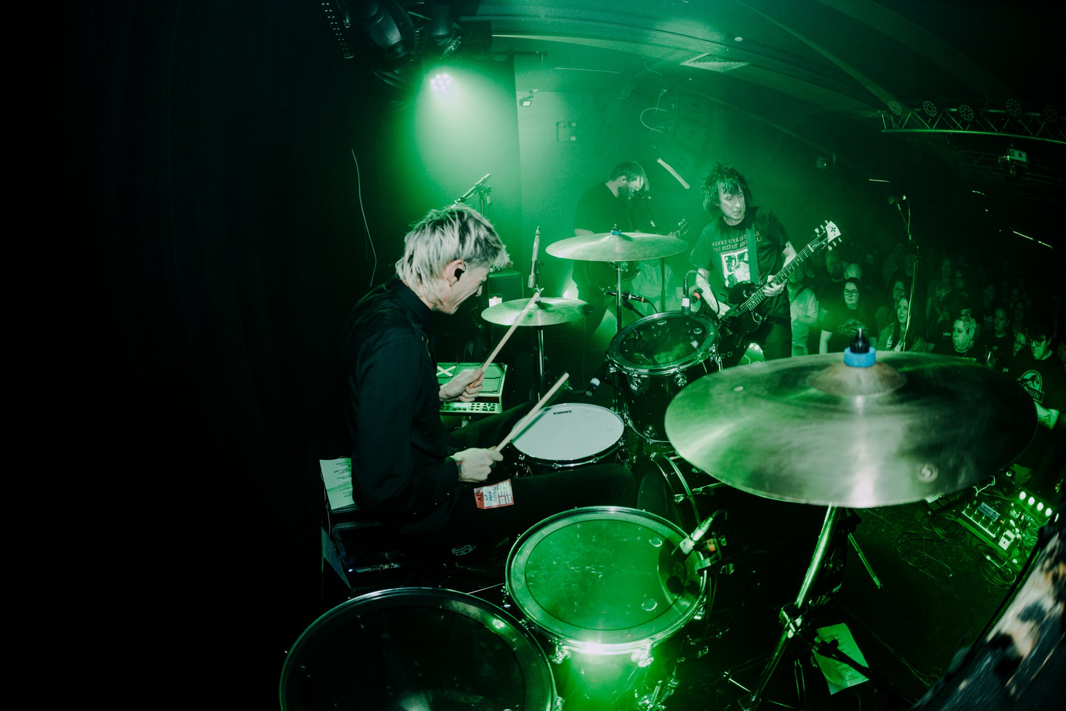 Band performing on stage with green lighting, featuring musicians playing drums, electric guitar, and keyboard, while audience watches in the background.