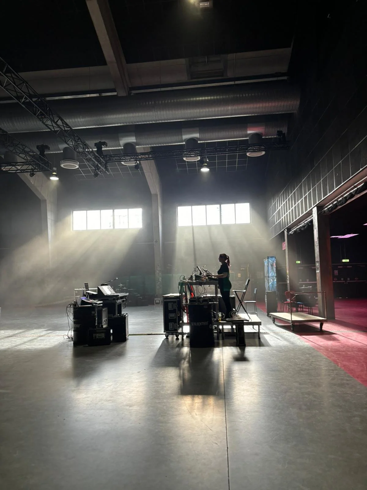 A performer wearing headphones operates electronic music equipment on a stage in a large, empty performance space with natural light coming through windows, and stage lighting overhead.