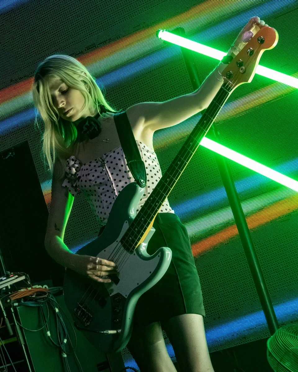 A woman playing a bass guitar on stage with green and multi-colored neon lights behind her.
