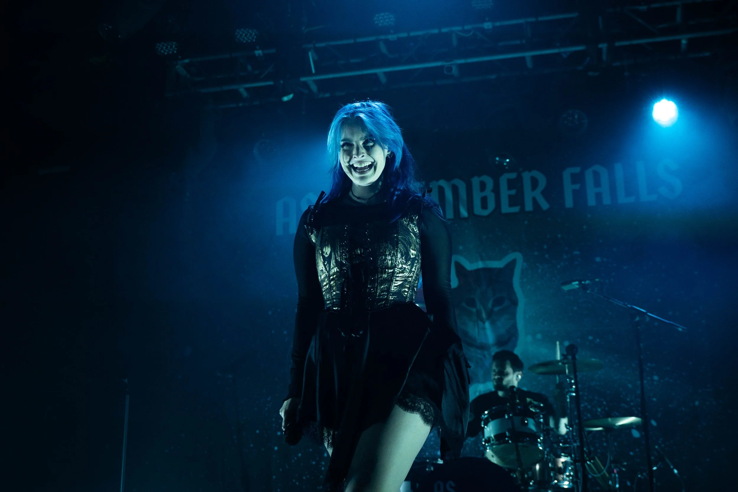 A female performer with blue hair and a metallic top on stage, smiling and looking at the camera, with a drummer in the background at a concert venue with a backdrop reading "Autumn OMSBER FALLS" and a cat logo.