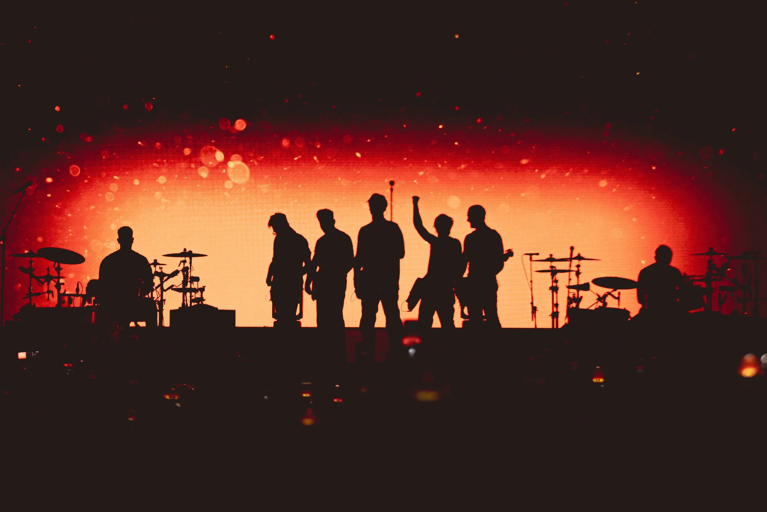 Silhouettes of a band performing on stage with drums and microphones against a red and orange background.