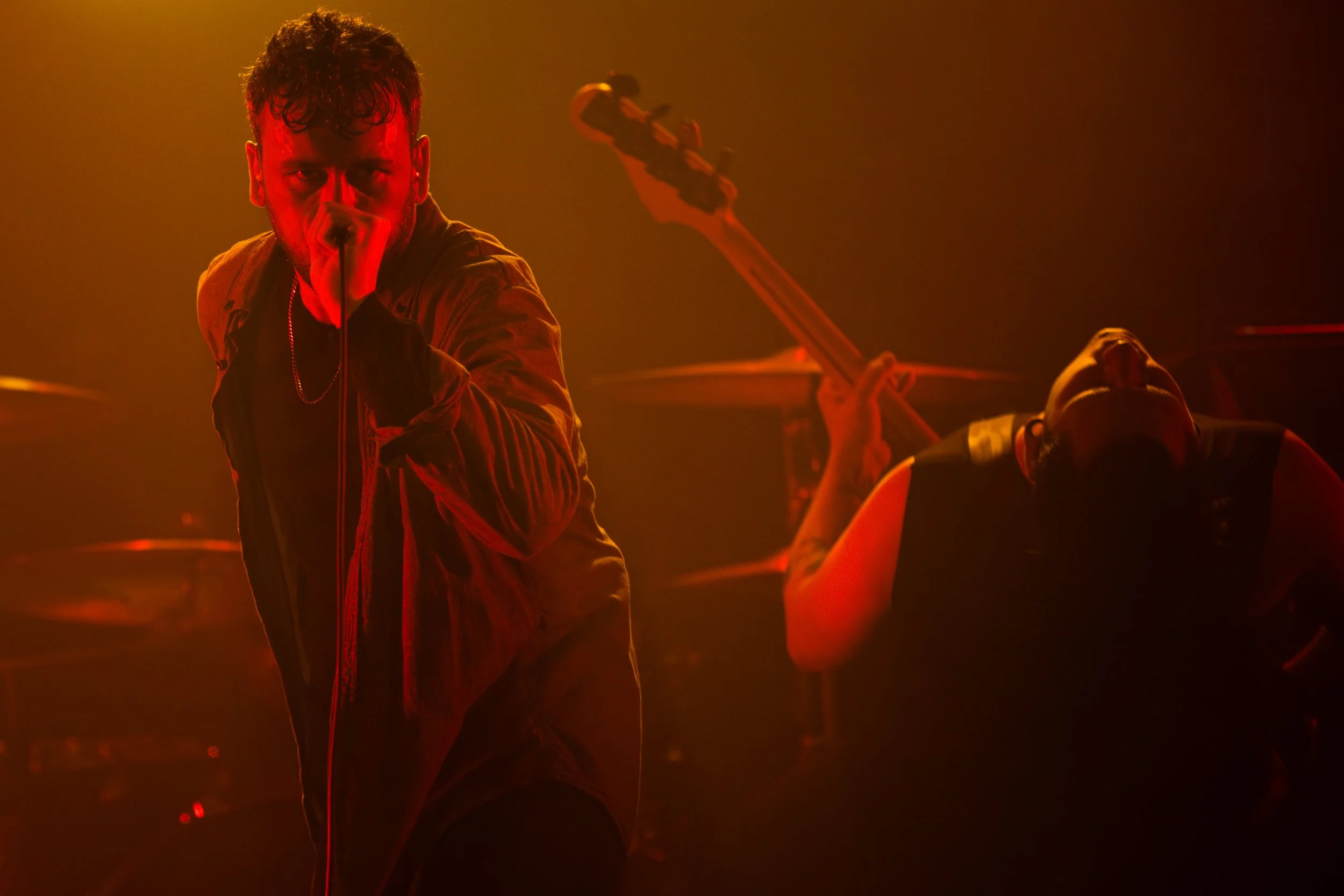 Male singer performs with microphone in left hand, looking intensely at the camera, with a drummer in the background playing drum set and a bassist playing bass guitar, all illuminated by red stage lighting.
