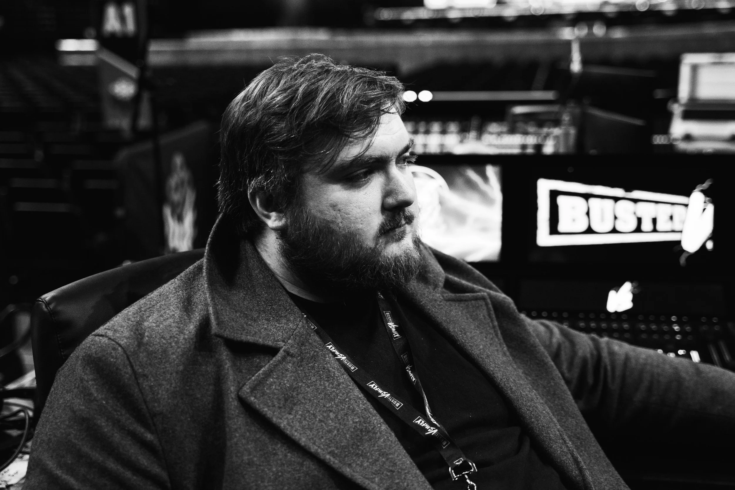 A man with a beard and long hair sitting indoors, wearing a coat and a lanyard, in front of a screen that says 'BUSTED'.