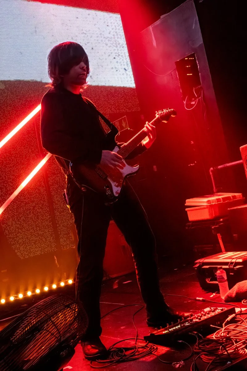 A person playing an electric guitar on stage with red lighting and a large screen in the background.