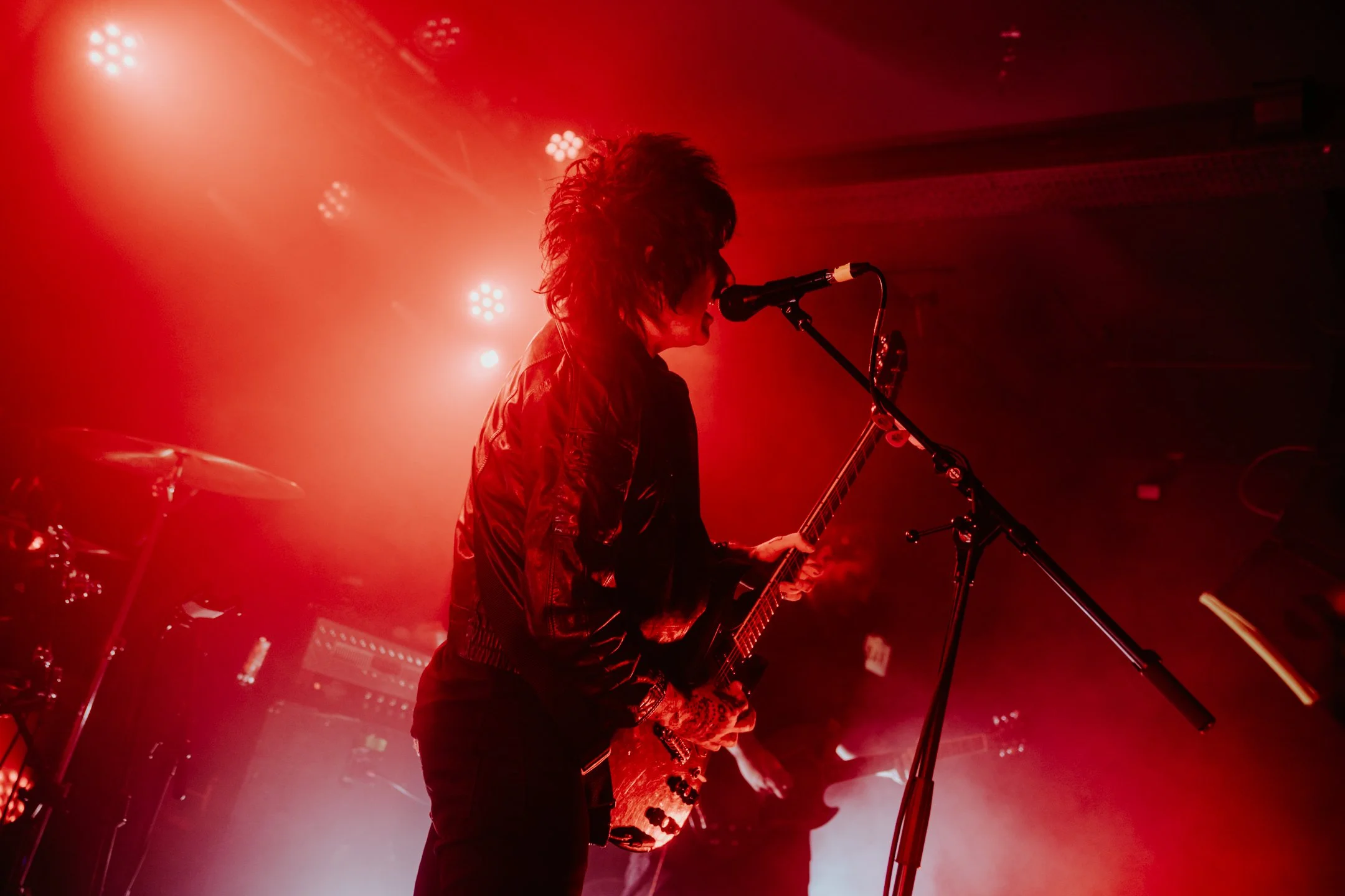 A musician performs on stage with red lighting, holding a guitar and singing into a microphone.