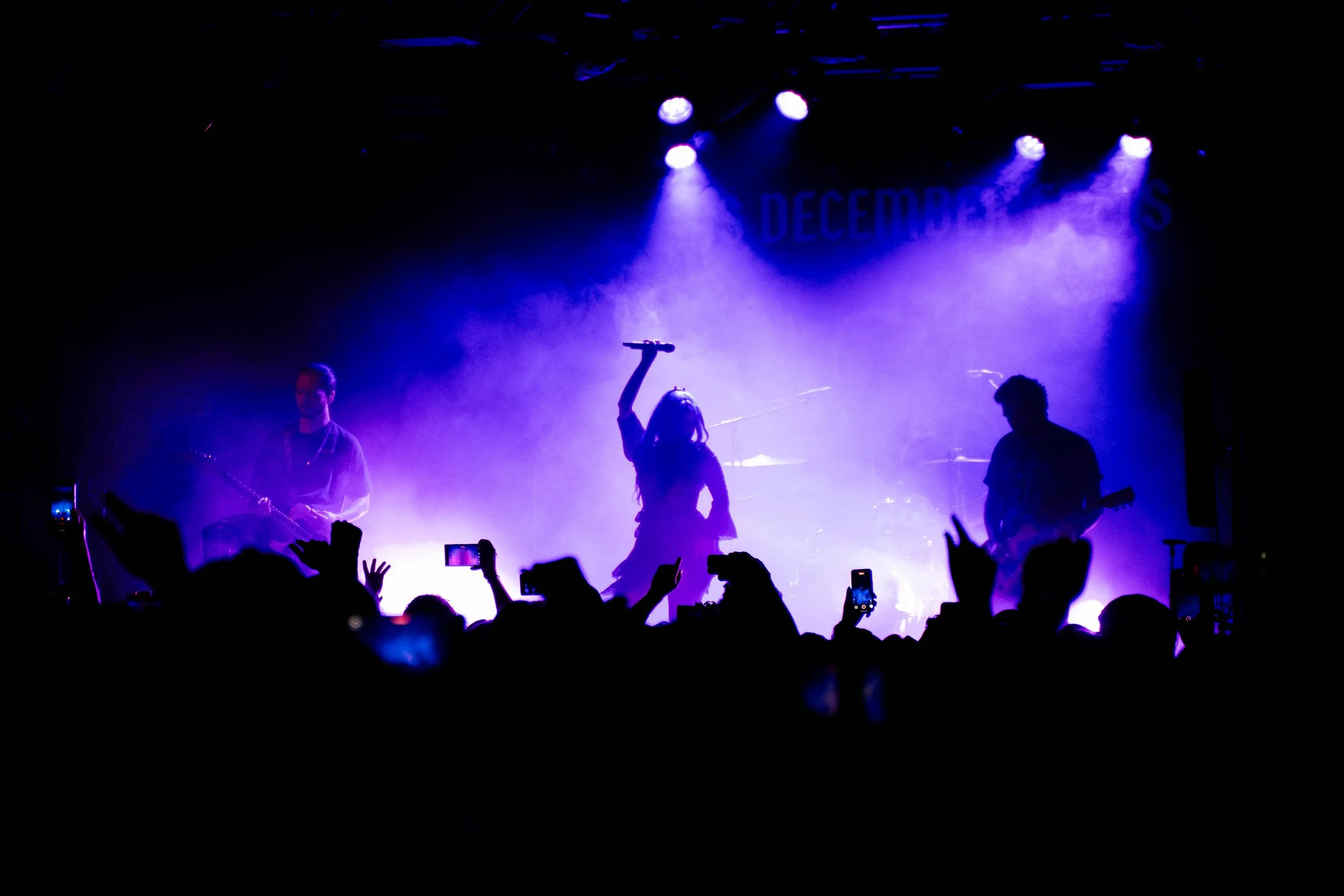 Silhouette of a female singer holding a microphone up with one hand on stage, flanked by two male guitarists on each side, with purple and blue lighting and an audience in the foreground taking photos.