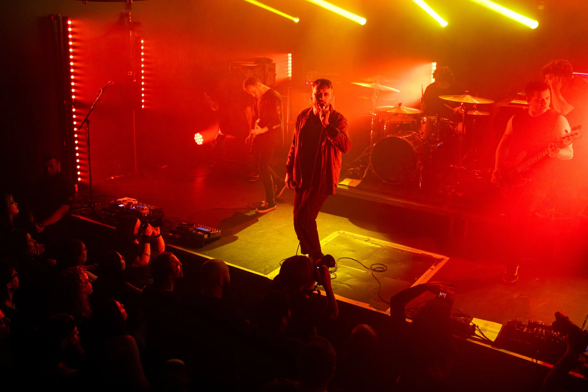 A live band performs on stage with yellow and red lighting, including a singer holding a microphone in the front, guitarists and a drummer in the background, and an audience watching from below.