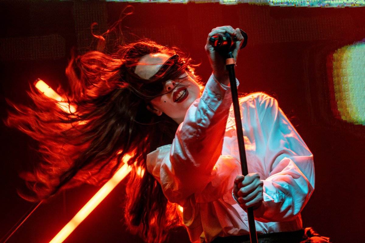 A woman with long hair singing into a microphone, wearing a white jacket and sunglasses, surrounded by red stage lighting.
