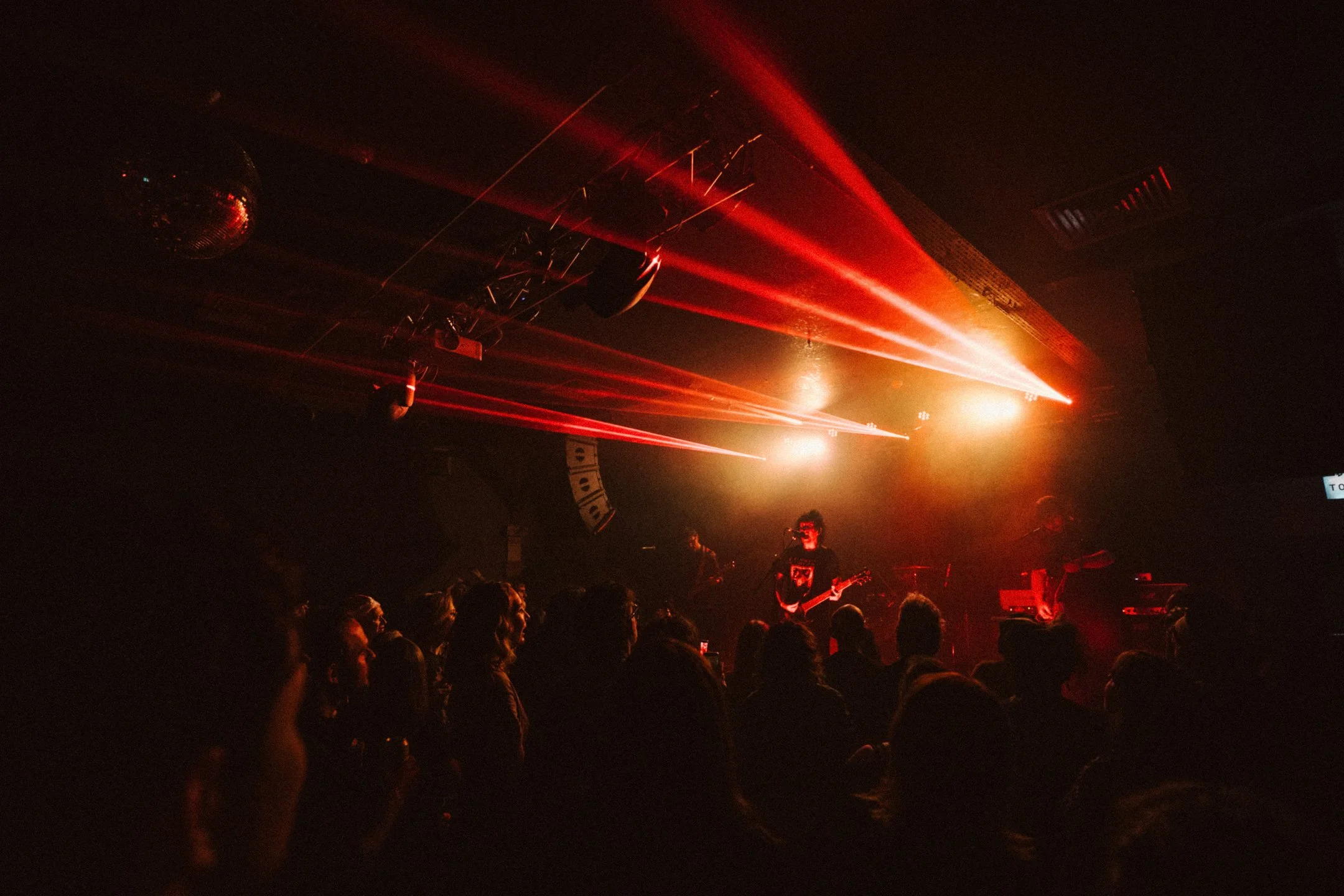 A live music concert with a band performing on stage, red laser lights illuminating the scene, and a dark audience in the foreground watching the performance.