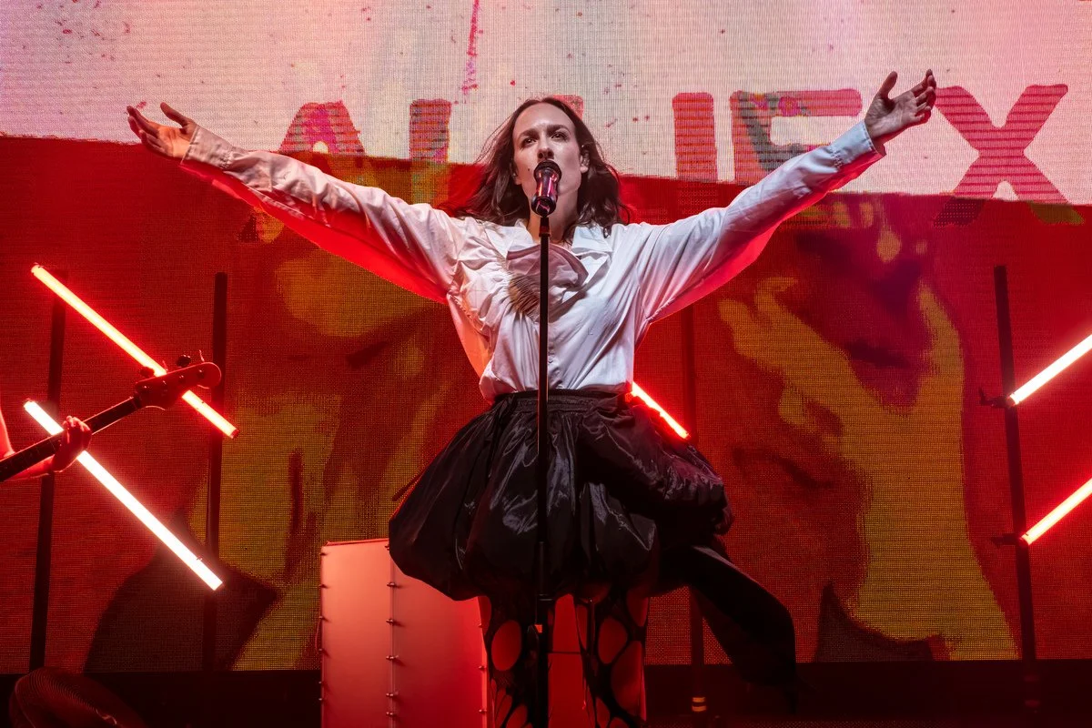 A woman in a white blouse and a black skirt performs on stage with her arms outstretched, singing into a microphone. The stage has red and black lighting with abstract LED light elements and a large screen in the background displaying the word 'ALIXY' with colorful visual effects.