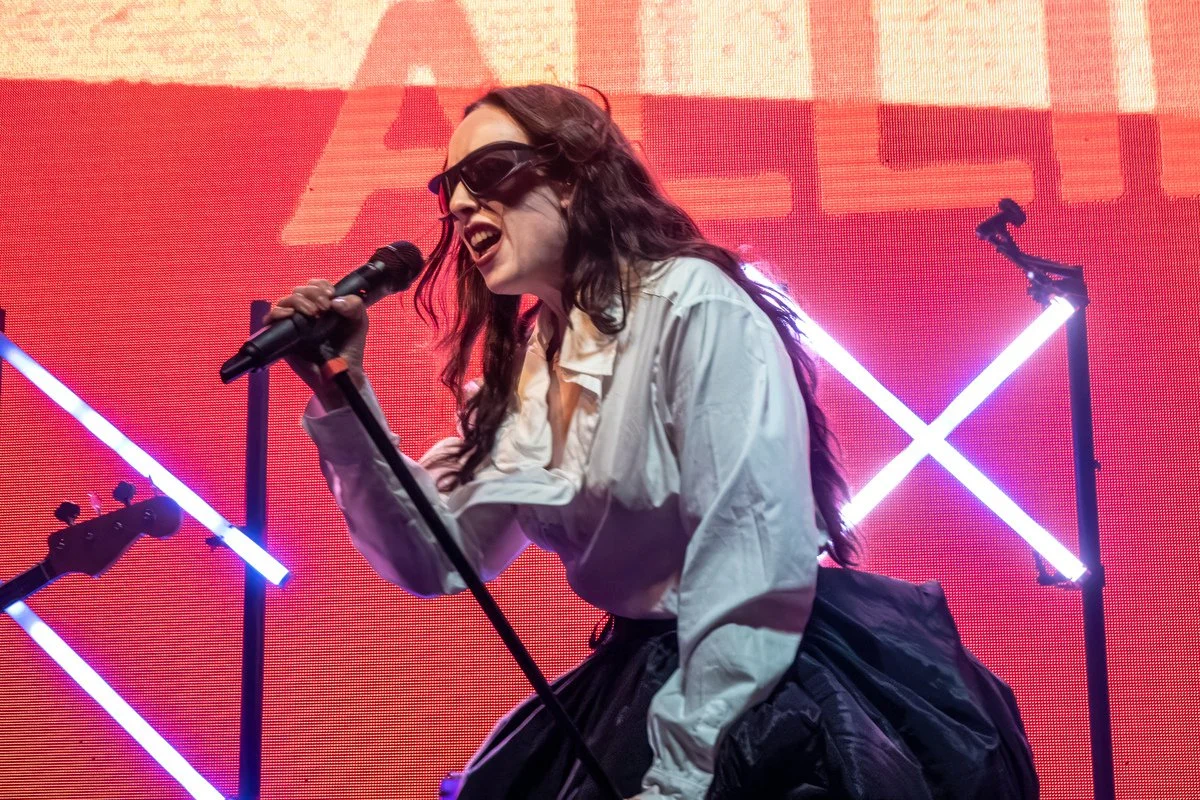 Female singer with long hair, sunglasses, white shirt, and black skirt, singing into a microphone on stage with neon lights and a red background.