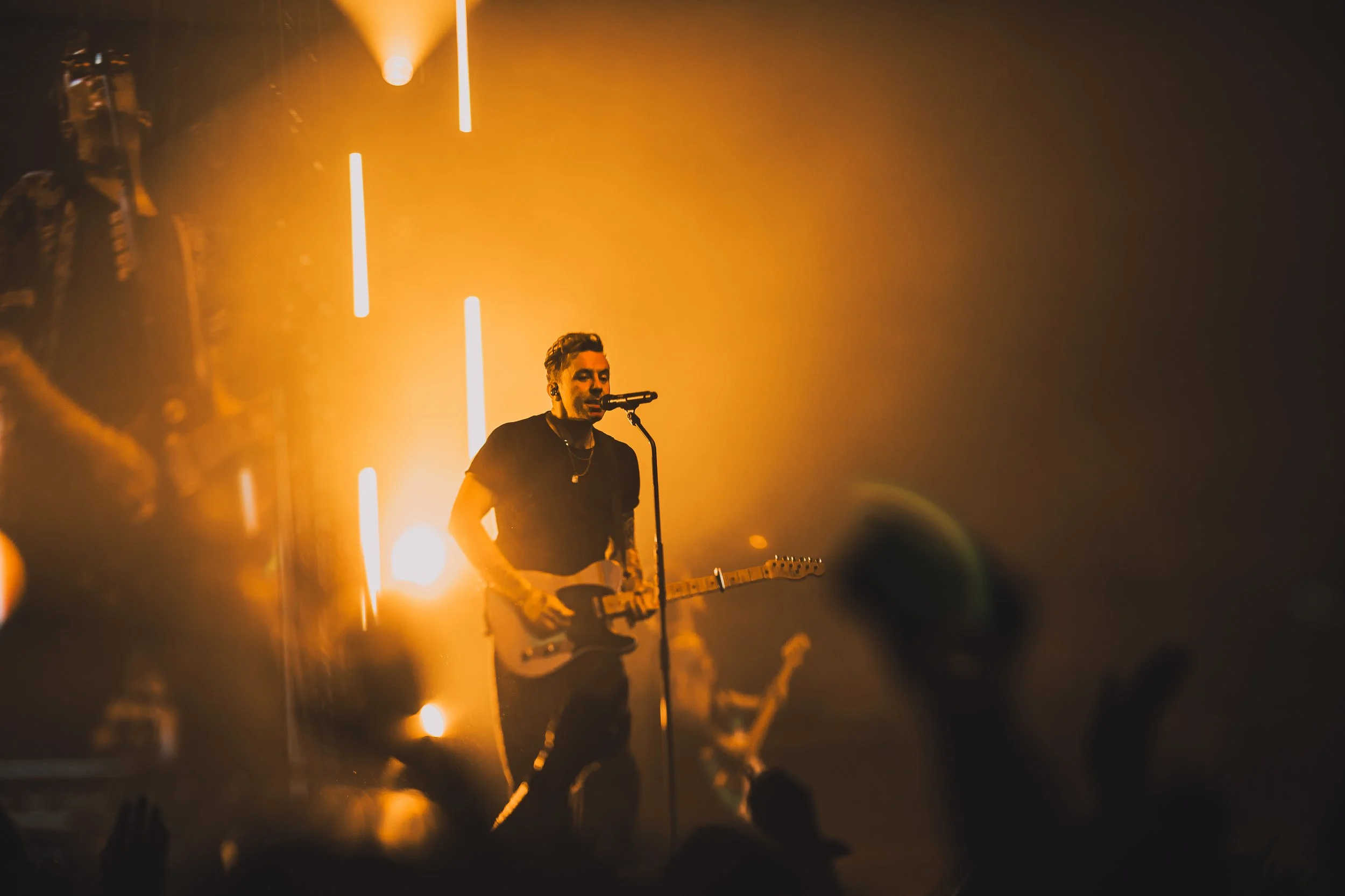 A male musician performing on stage with an electric guitar, performing under orange stage lights, and an audience with raised hands.