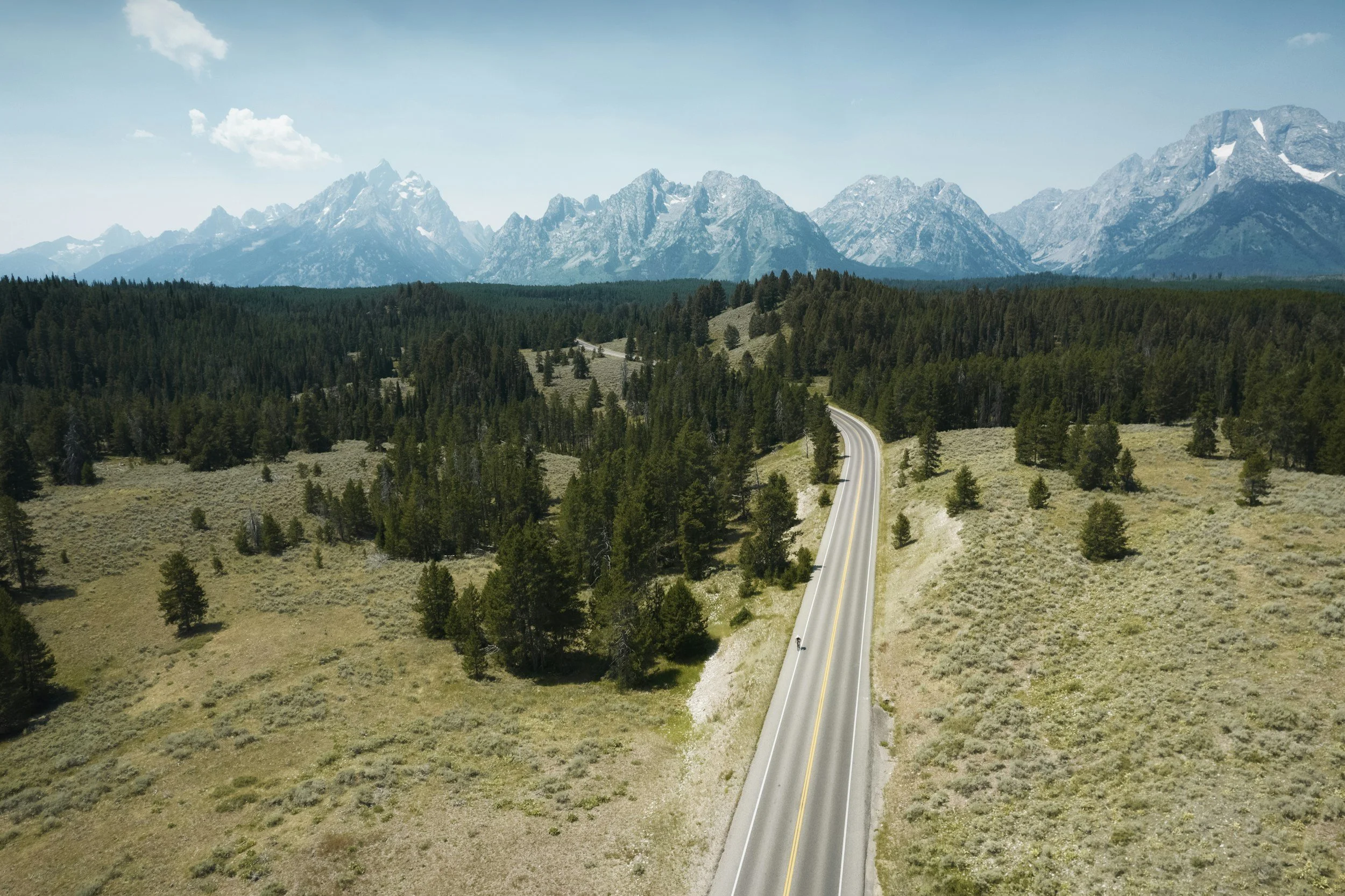 A winding road cuts through a grassy landscape with scattered trees, leading towards distant mountain ranges under a blue sky with a few clouds.