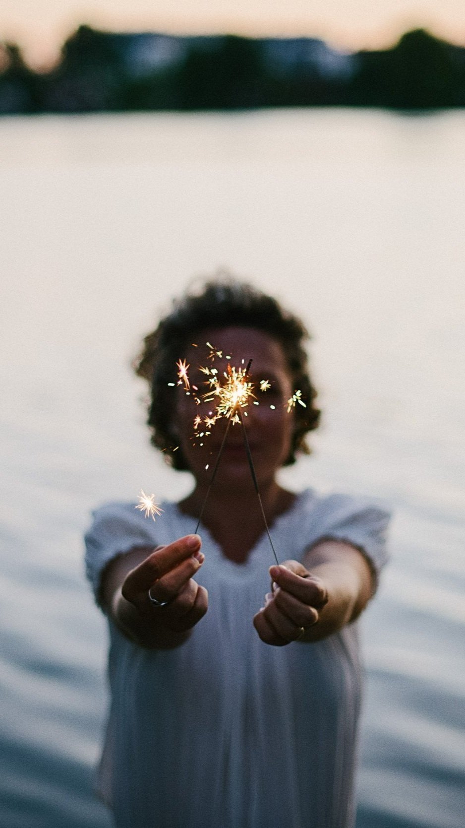 A woman standing near water at dusk holding a lit sparkler towards the camera, with the river and distant land in the background.