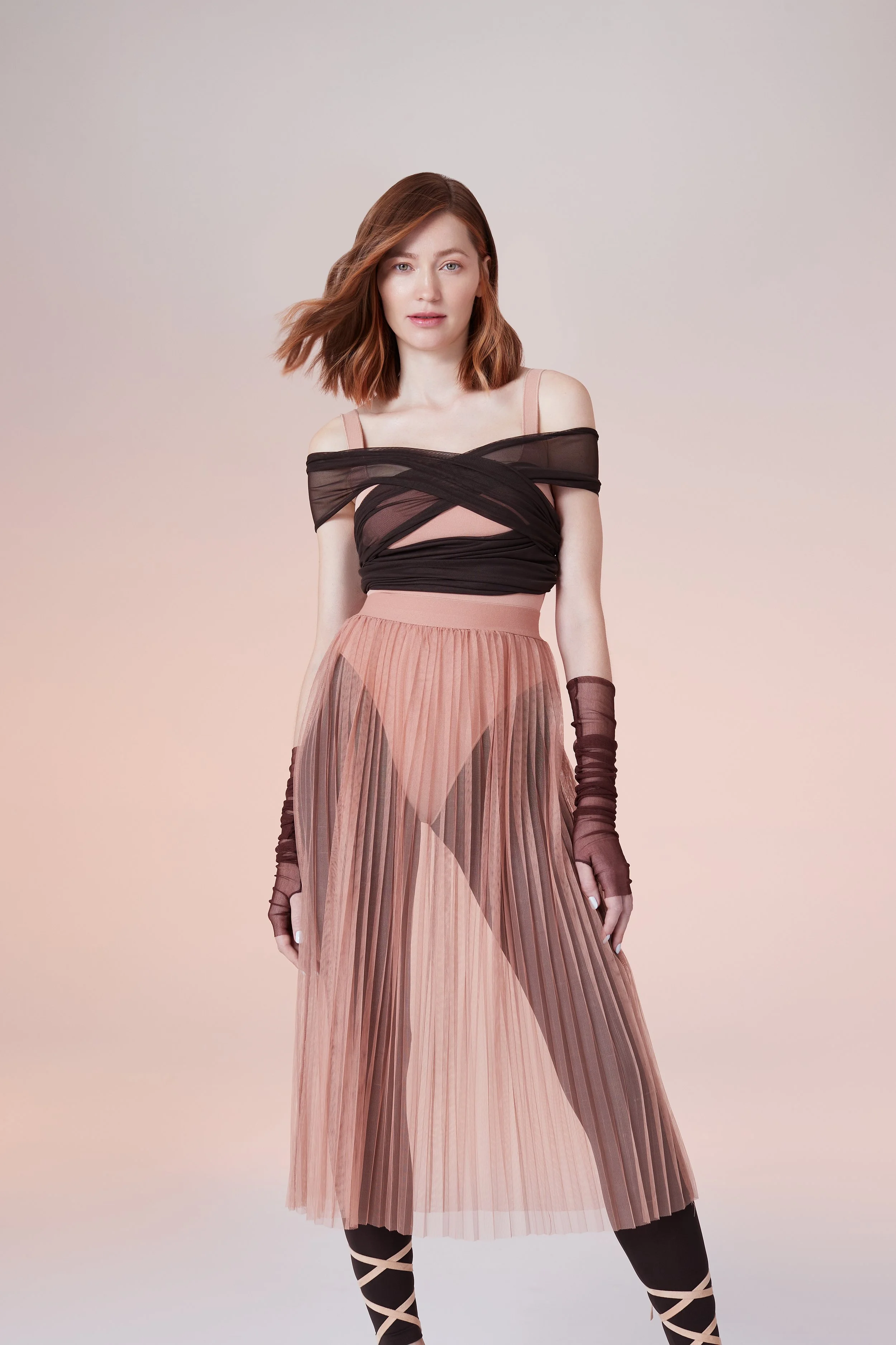 A woman with red hair wearing a pink and black dress with sheer fabric and pleats, along with arm sleeves and crisscross leggings.