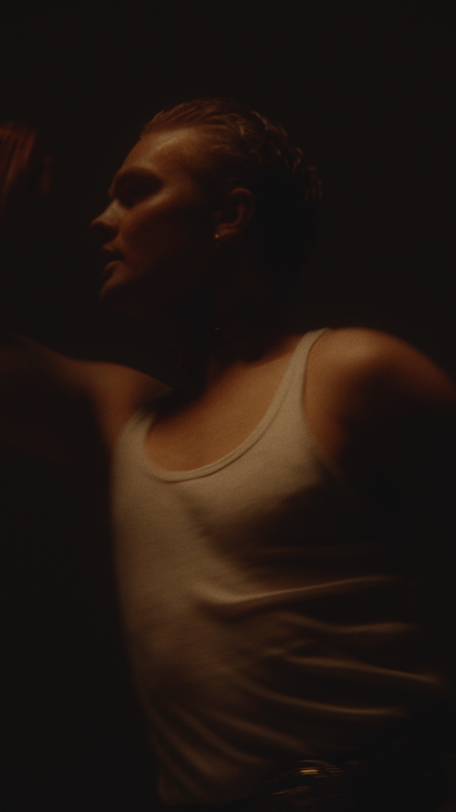 A woman with short hair styled back, wearing a white tank top, looking to her left in a dimly lit setting.