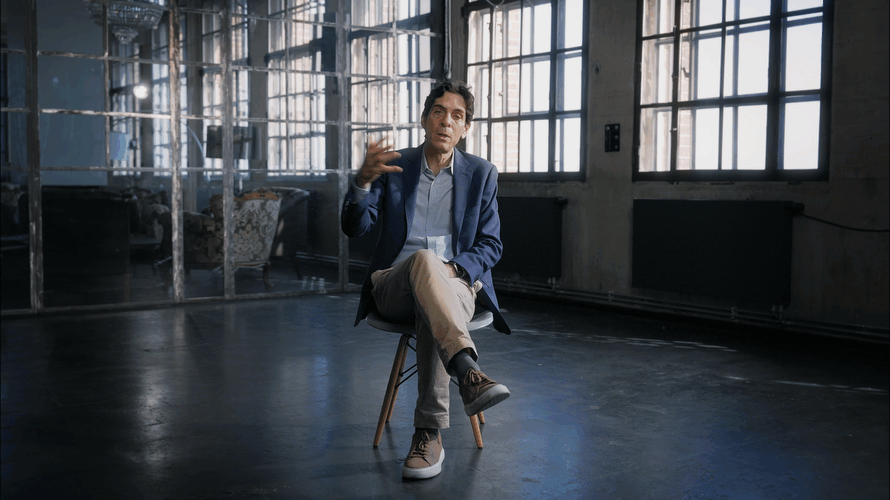 A man sitting on a chair in an industrial-style room with large windows, wearing a blue blazer, light-colored pants, and sneakers, speaking or explaining something.