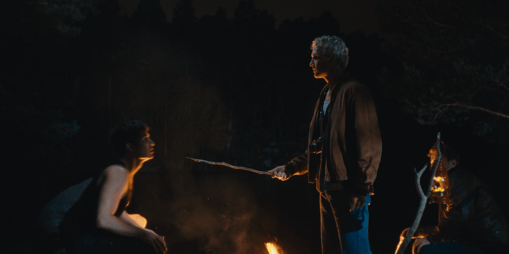A person with gray hair holding a stick confronts a woman sitting by a campfire at night. Two other people are visible in the background, one holding a lantern.