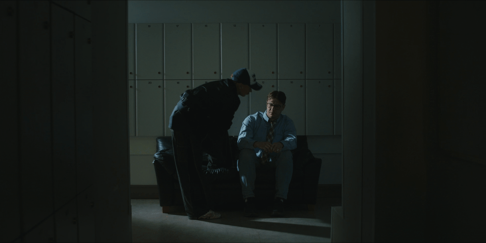 A man wearing glasses and a blue shirt sits on a black sofa, while another person leans over him in a dimly lit room with lockers in the background.
