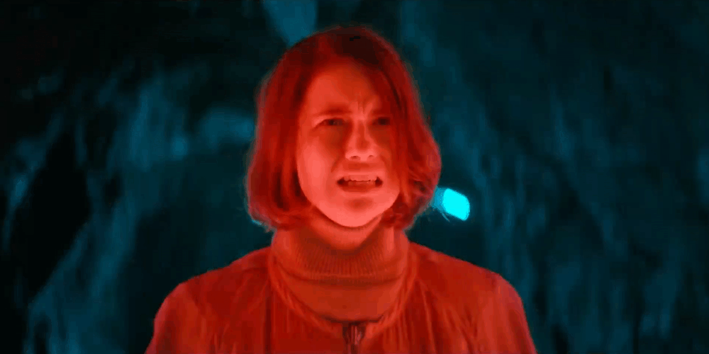 A woman with short red hair looks distressed inside a dark cave with glowing blue light.