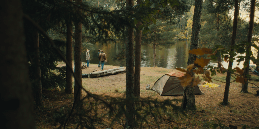 People camping by a river in a forested area with a tent and trees around.
