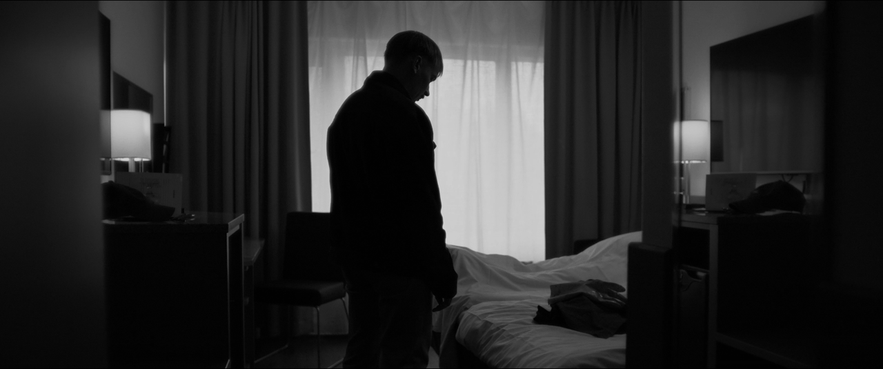 Silhouette of a man standing beside an unmade bed in a hotel room, backlit by a window with curtains.