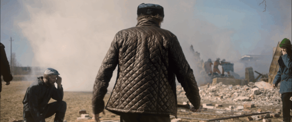 A person in a quilted jacket walking through a scene of destruction with smoke, with other individuals nearby amidst rubble.