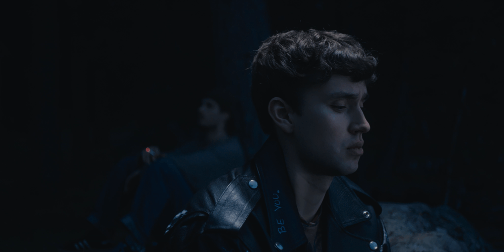 A young man with dark curly hair, wearing a black leather jacket, stands in a dark, moody landscape with a somber expression. Another person is faintly visible in the background with blurred features.