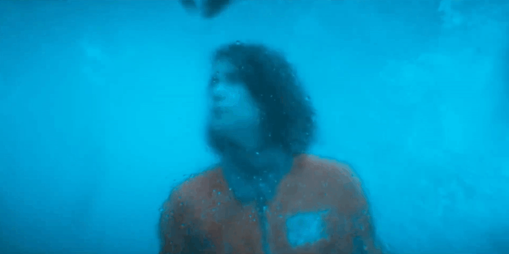 A woman with curly hair and wearing an orange jacket is underwater, looking at the surface with bubbles surrounding her.