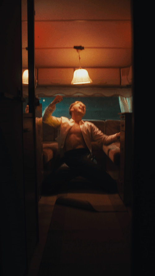 A person in a white unbuttoned shirt and dark pants strikes a confident pose inside a small, dimly lit room with a ceiling light and a window with blue curtains.