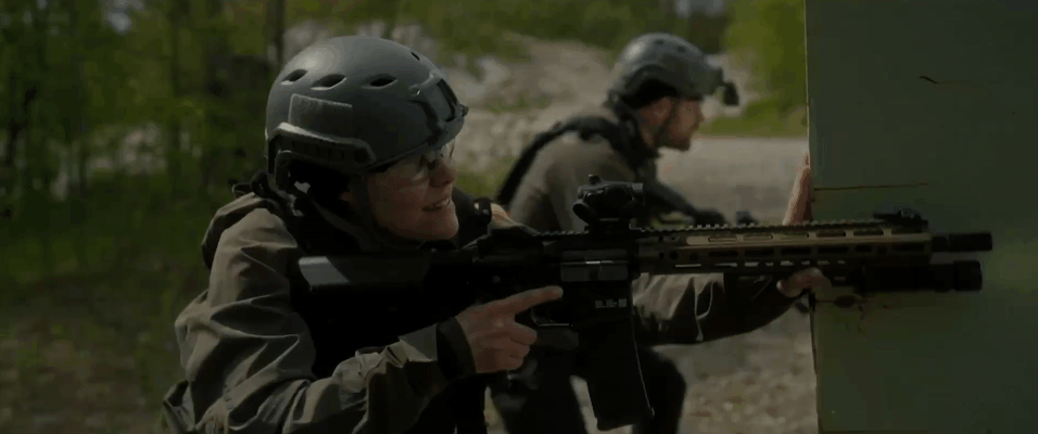 Two soldiers wearing helmets and tactical gear firing rifles behind cover in a wooded area.