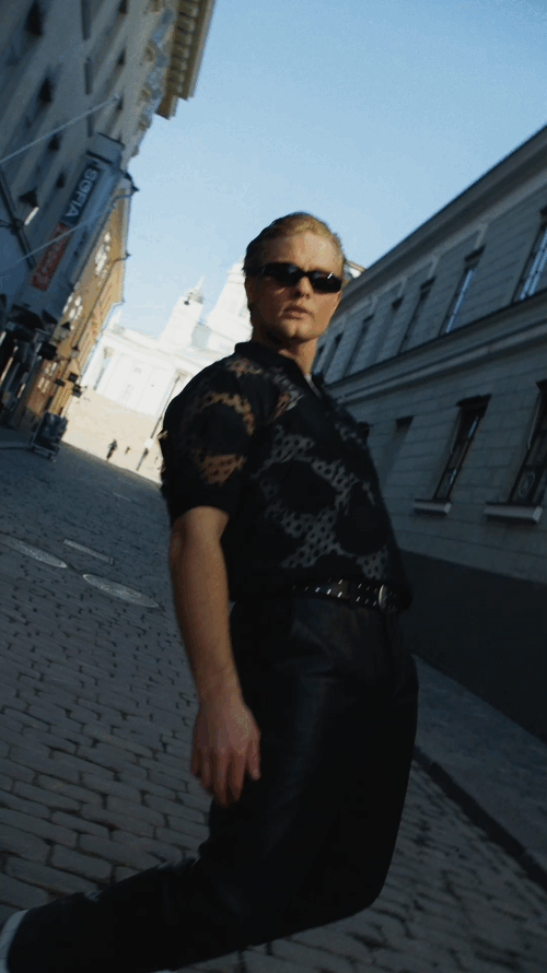 Person standing on a cobblestone street in a city, wearing dark sunglasses, a black lace shirt, and black pants, with historic buildings in the background.