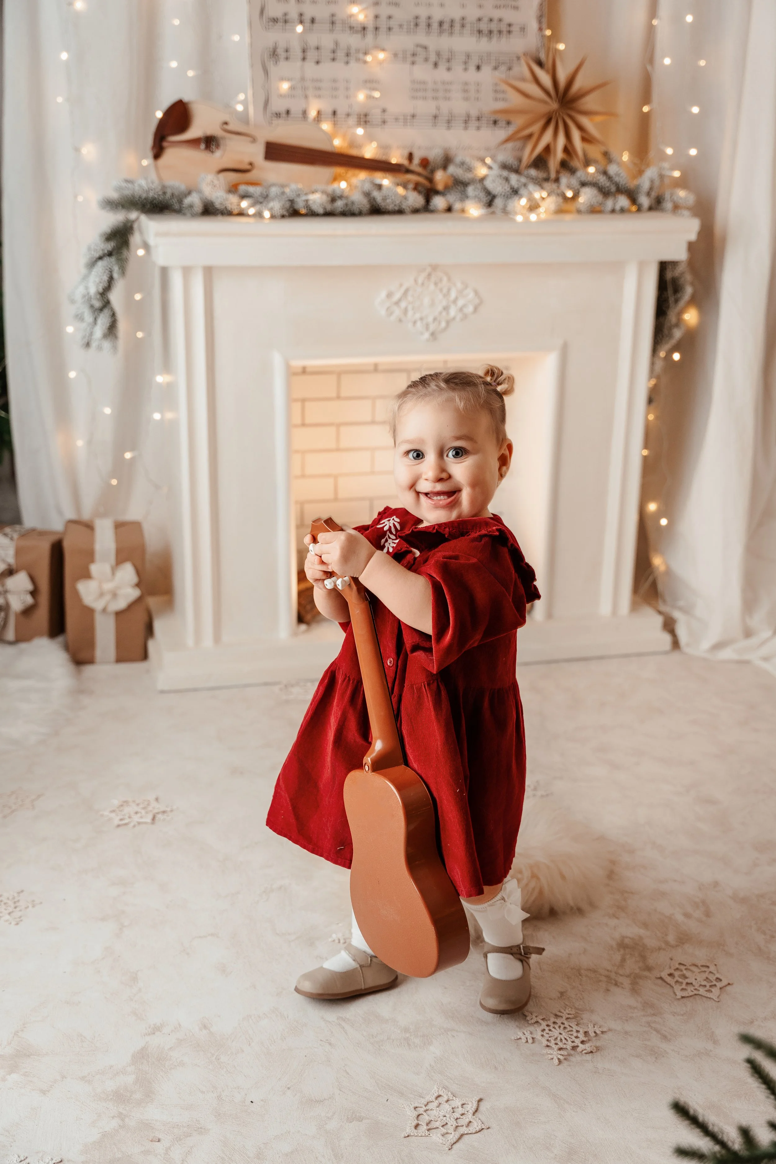 Niña sonriendo en una sala decorada con motivos navideños, sosteniendo una guitarra de juguete, con una chimenea decorada con luces y adornos, y con regalos debajo.