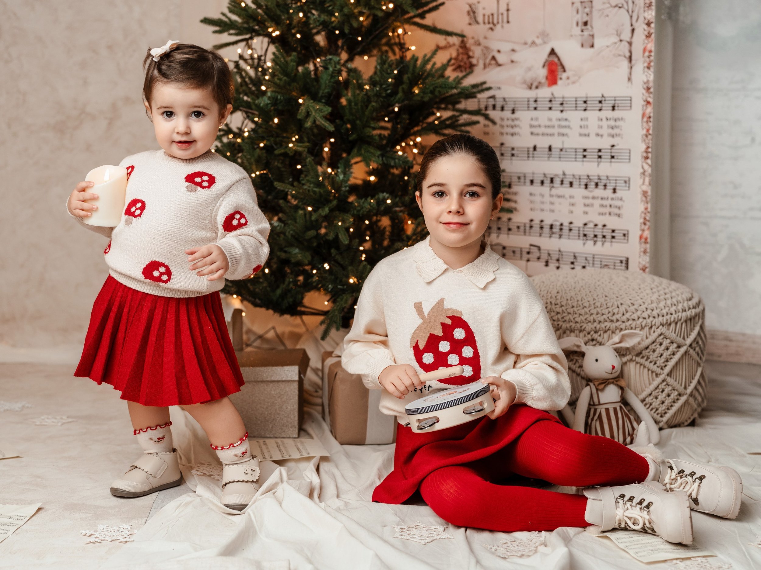 Dos niñas en Navidad, una con suéter con dibujos de hongos y falda roja, la otra con suéter con calabaza y pantalones rojos, en una sala decorada con árbol de Navidad y música navideña en la pared.