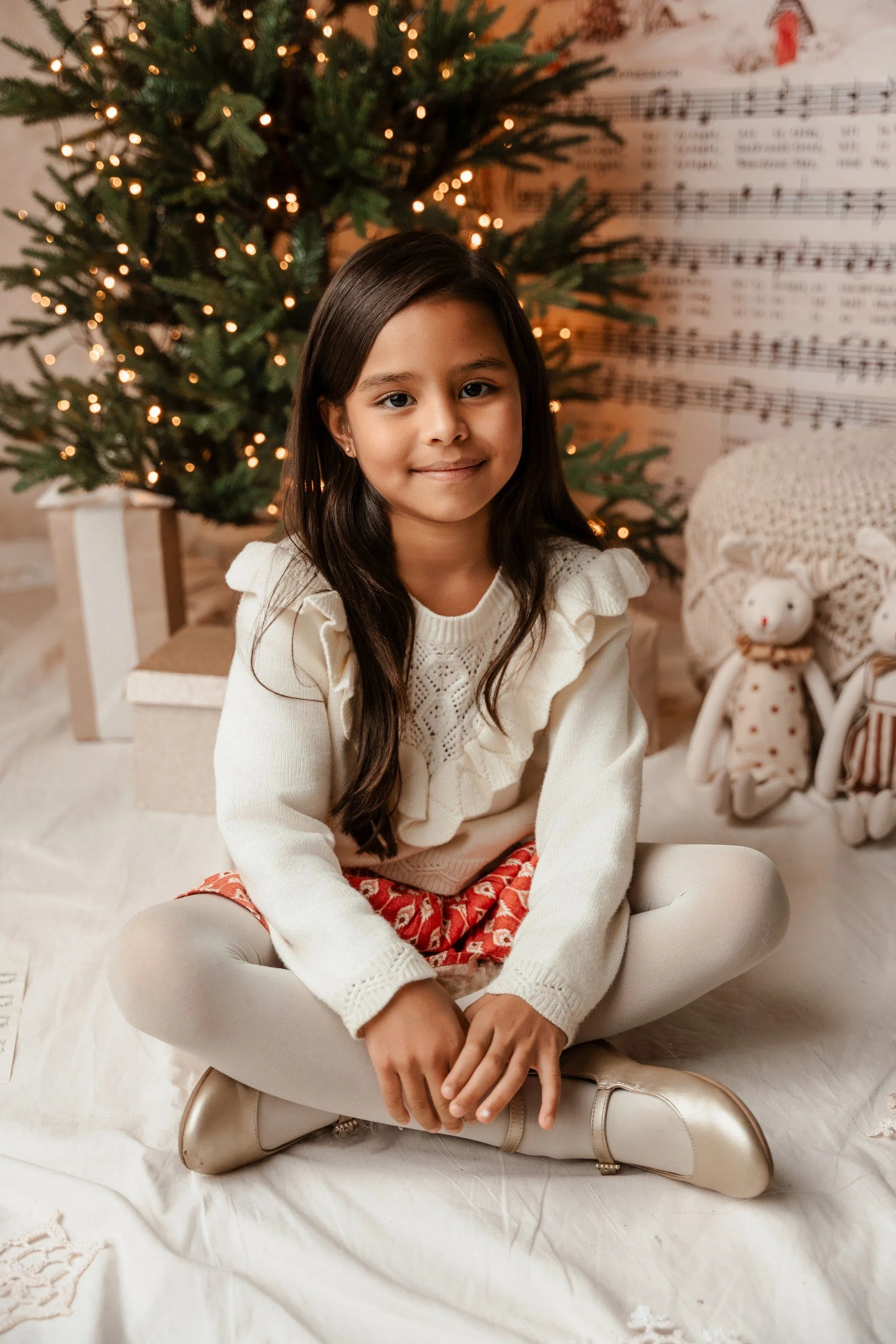 Niña sonriendo sentada en el suelo con árbol de Navidad decorado y música de villancicos en el fondo, rodeada de decoración navideña.