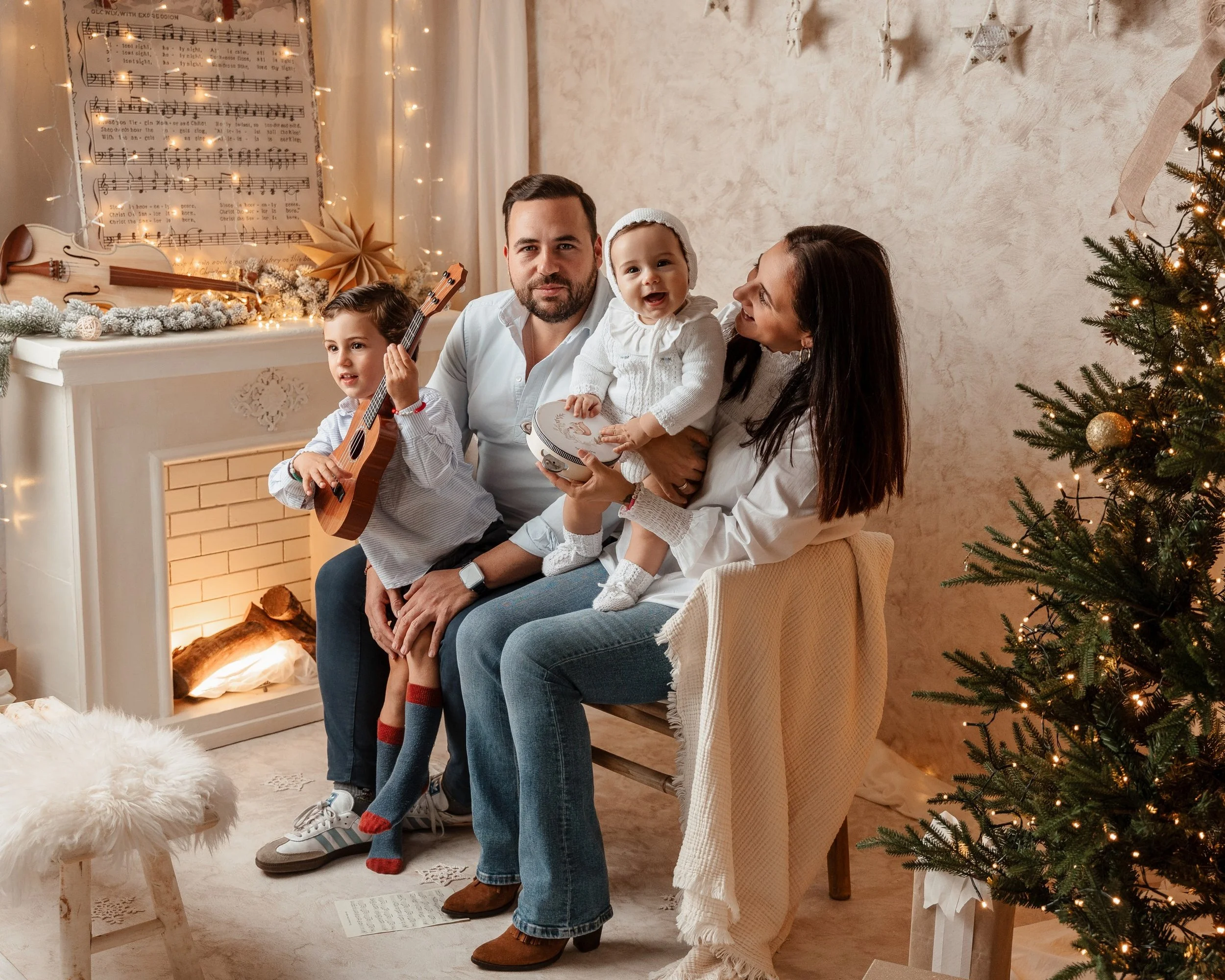 Una familia de cuatro personas celebra la Navidad en una sala decorada con un árbol de Navidad iluminado, estrellas blancas en la pared, una figura musical y luces decorativas. La madre sostiene a una niña pequeña que tiene un tambor, el padre sostie
