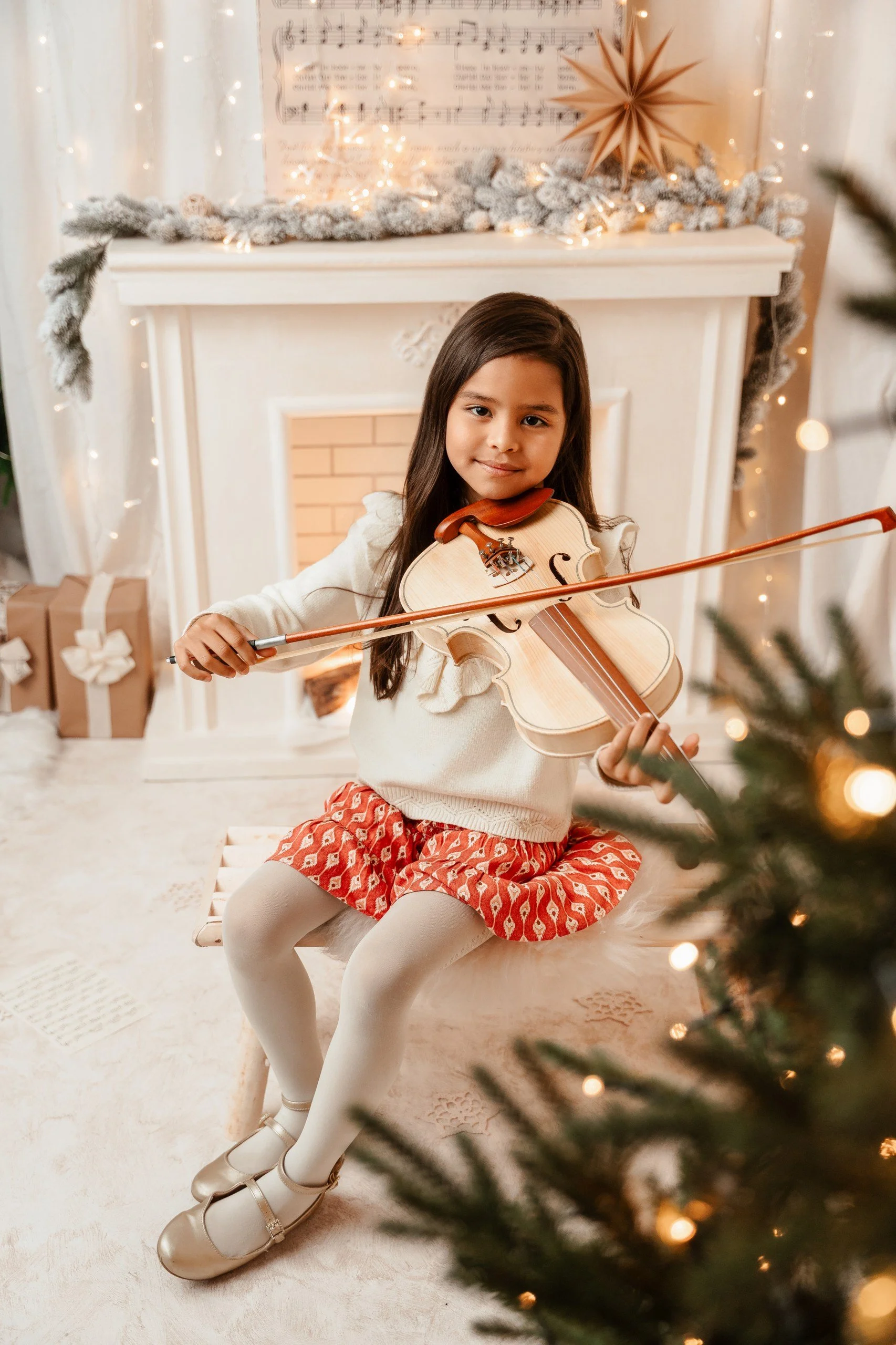 Niña tocando el violín en un decorado navideño con árbol de Navidad, regalos y adornos en el fondo.