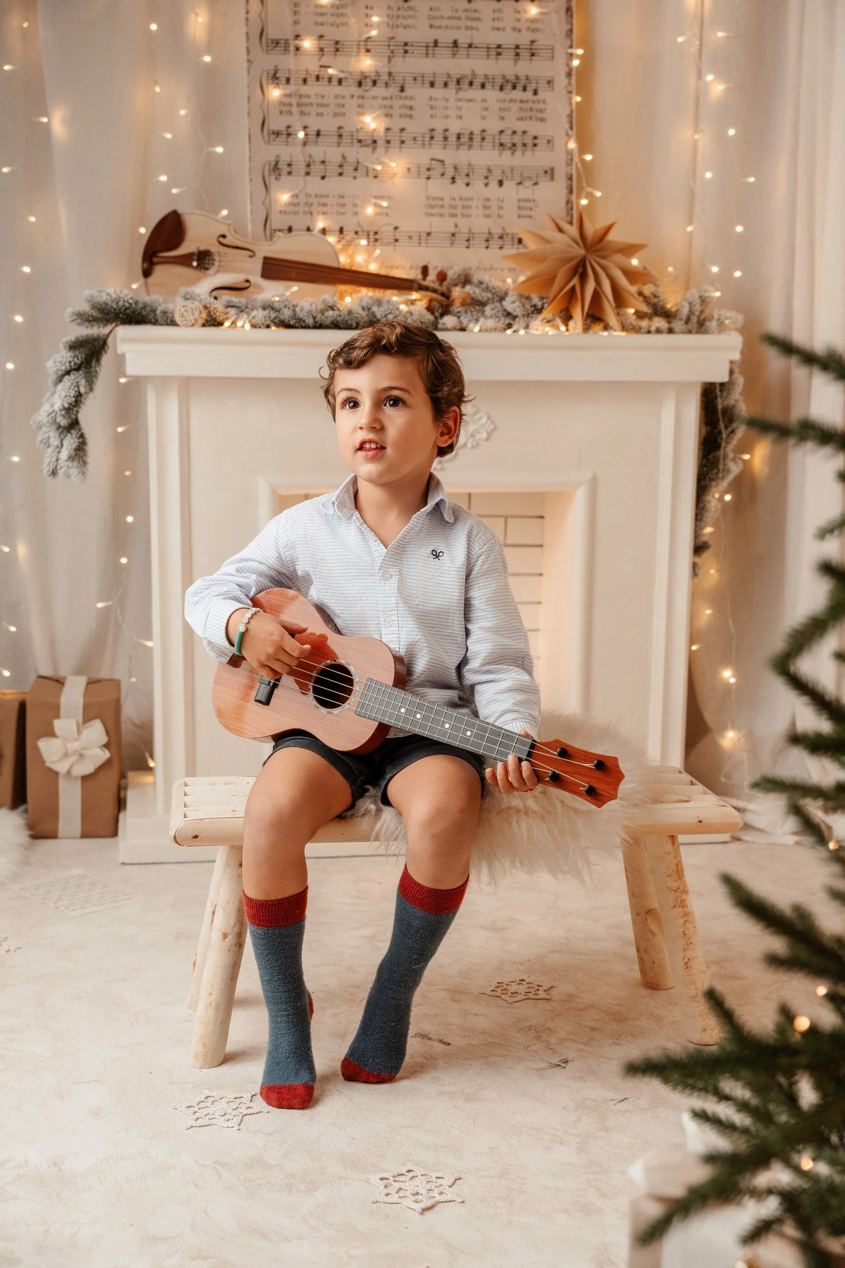 Niño sentado en una banca de madera sosteniendo una guitarra pequeña en un ambiente decorado con luces de Navidad, un árbol de Navidad parcialmente visible en la esquina derecha y decoraciones navideñas en el fondo.